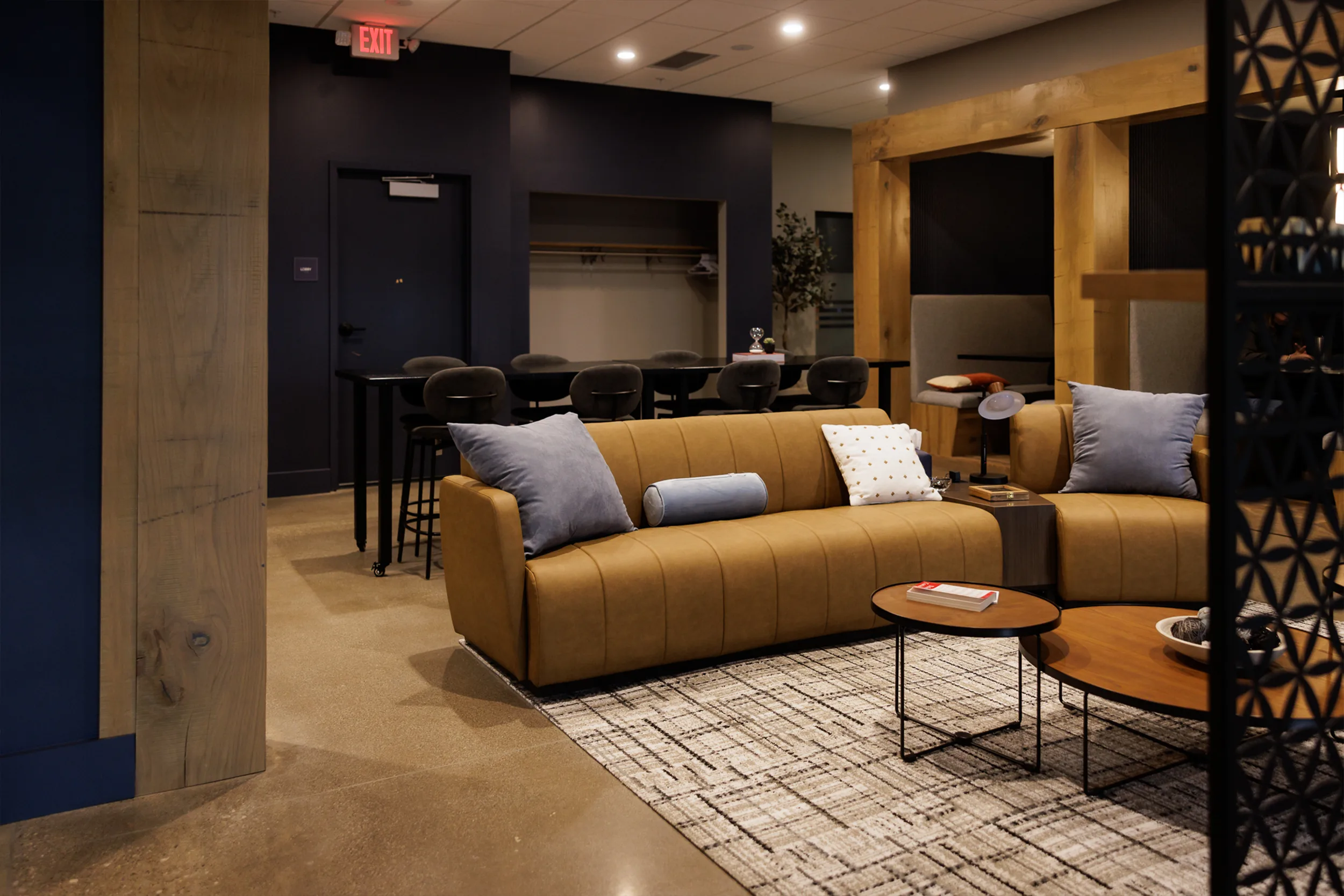 Modern lounge interior with cozy brown couch, assorted pillows, round tables, and high seating area. Soft lighting and contemporary decor create inviting atmosphere.