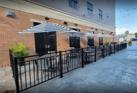Outdoor patio area with black tables and chairs under striped umbrellas, surrounded by a black fence.