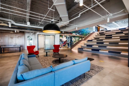 Modern office lobby featuring a stylish blue sofa, red accent chairs, and a round coffee table, complemented by a vibrant mural and illuminated staircase. The space showcases contemporary design elements and a welcoming atmosphere.