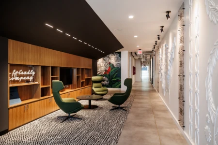 Modern office interior featuring a stylish hallway with green lounge chairs, a wooden shelving unit, and decorative wall art. The space is well-lit with contemporary lighting and has a sleek, minimalist design, creating an inviting atmosphere for collaboration and relaxation.