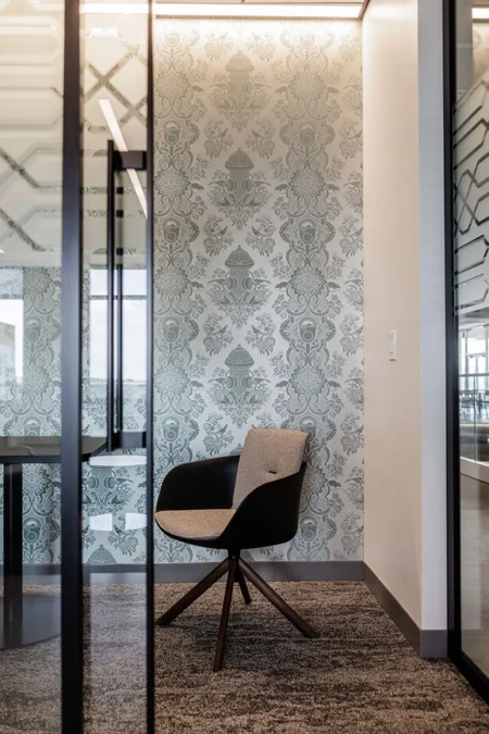 Modern office space featuring a stylish black and gray chair against an elegant patterned wallpaper, with glass walls and a contemporary table in the background, creating a sophisticated work environment.