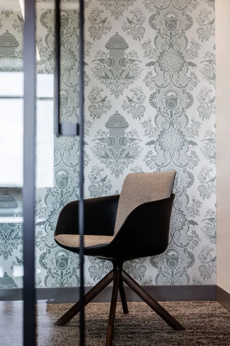 A modern chair rests on carpeting, near ornately patterned wallpaper featuring detailed designs, behind a part-glass partition in a stylish interior space.