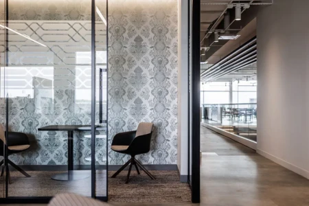 Modern office interior featuring a glass-walled meeting room with stylish chairs and a decorative wallpaper. The image showcases a sleek, open hallway leading to a spacious collaborative area with natural light.