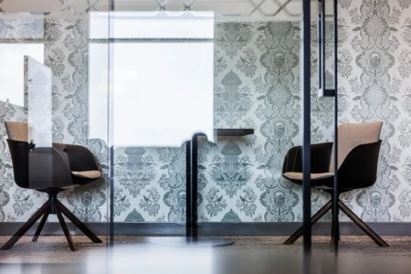 Modern office meeting space featuring two stylish chairs and a small table, surrounded by decorative wallpaper with intricate patterns, illuminated by natural light.