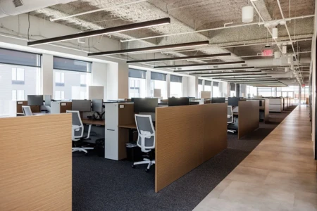 Modern open-plan office with wooden cubicles, white chairs, and computers. Large windows provide natural light; exposed ceiling displays industrial design. Quiet, organized workspace.