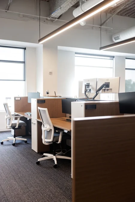 Modern office workspace featuring ergonomic chairs and dual monitor setups, designed for productivity and collaboration, with natural light from large windows and stylish wooden partitions.