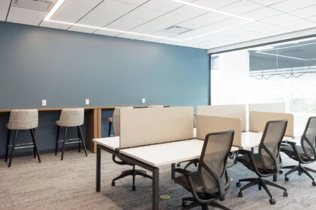 A modern, spacious office with individual workstations, swivel chairs, a wooden counter with barstools, and a large window providing natural light.