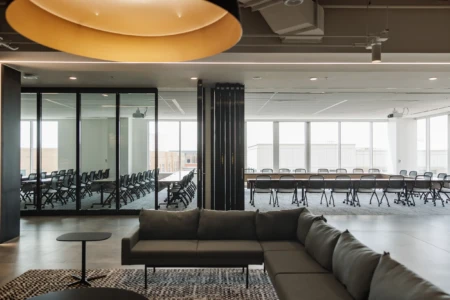 Modern office interior featuring a stylish lounge area with a gray sectional sofa and a small black table, overlooking a large conference room with rows of black chairs and a long wooden table, illuminated by natural light from large windows.