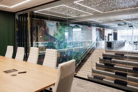 Modern office conference room featuring a large wooden table with white ergonomic chairs, glass walls showcasing vibrant artwork, and a stylish staircase leading to a communal area with a kitchen and seating.