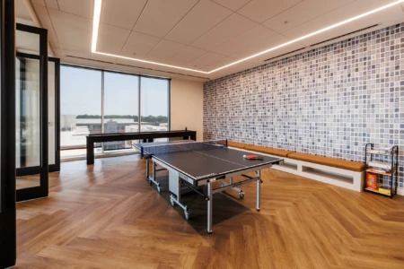 Modern office game room featuring a ping pong table, large windows with a scenic view, and a stylish wall adorned with patterned tiles. The space includes a comfortable bench and a shelf with games, emphasizing a relaxed work environment.