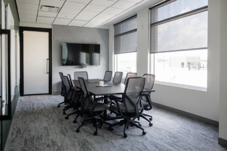 Modern conference room featuring a large black table surrounded by ergonomic mesh chairs, a wall-mounted TV, and large windows with roller shades. The room is designed for meetings and collaboration, with a neutral color palette and carpeted flooring.