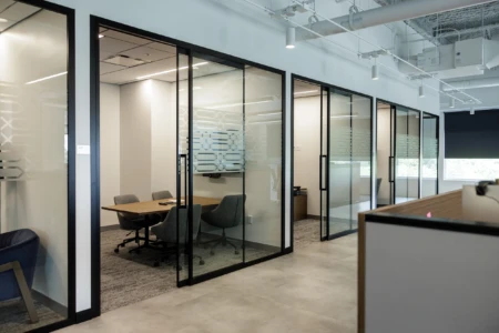 Modern office interior featuring glass-walled meeting rooms with wooden tables and ergonomic chairs, designed for collaboration and productivity. The space includes contemporary decor and ample natural light.