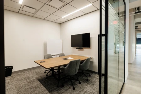 Modern conference room featuring a wooden table, four ergonomic chairs, a wall-mounted TV, and a whiteboard, with large glass doors providing a view of the adjacent area.