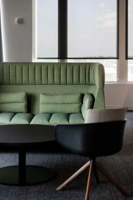 Modern green upholstered sofa with vertical tufting and two small cushions, paired with a sleek black chair on a textured gray carpet, set against large windows. Ideal for contemporary office or lounge spaces.