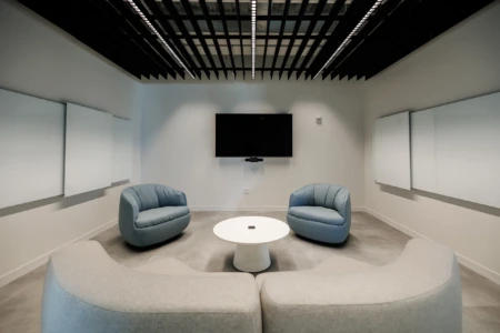 Modern meeting room featuring comfortable blue seating, a round white table, and a large wall-mounted television. The space includes writable whiteboards and a stylish black ceiling design, ideal for collaborative discussions and presentations.