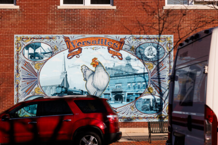 Mural depicting the town of Versailles, Ohio, featuring a large chicken illustration and historical imagery, set against a brick background. A red vehicle is partially visible in the foreground.