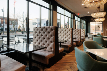 Stylish restaurant interior featuring plush tufted booths, modern furnishings, and large windows offering a view of the street outside.