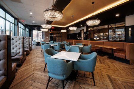 Modern restaurant interior featuring elegant chandeliers, cozy seating areas, and a warm wooden floor. The space includes stylish blue chairs and cushioned booths, creating an inviting atmosphere for dining. Large windows allow natural light to illuminate the contemporary design.
