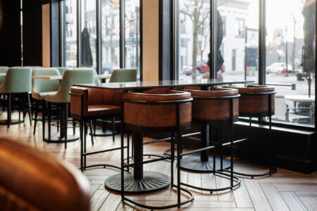 Modern restaurant interior featuring stylish brown leather bar stools and mint green chairs, with large windows allowing natural light and a view of the street outside. The elegant wooden flooring complements the contemporary design.