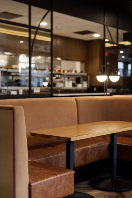Cozy restaurant booth with brown leather seats and wooden table. Open kitchen visible behind glass, with softly lit ambiance and modern decor.