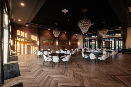 Elegant event space featuring round tables with white chairs, stylish chandeliers, and a warm brick accent wall, designed for gatherings and celebrations. Natural light streams in through large windows, enhancing the inviting atmosphere.