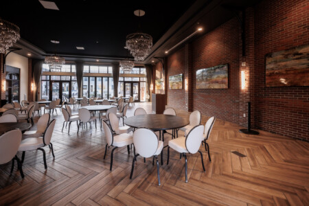 Interior view of a modern dining space featuring elegant round tables with white chairs, hardwood herringbone flooring, and large windows allowing natural light. The walls are adorned with contemporary artwork, and stylish chandeliers provide ambient lighting, creating a sophisticated atmosphere for dining or events.