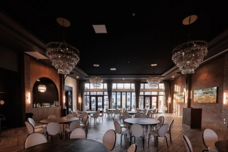 Elegant event space featuring round tables and white chairs, adorned with crystal chandeliers. Large windows allow natural light to fill the room, highlighting the stylish decor of exposed brick and wooden accents, ideal for gatherings and celebrations.