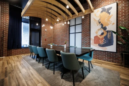 Modern conference room with wooden ceiling design, brick walls, abstract artwork, long table, blue chairs, large window, and hanging lights.
