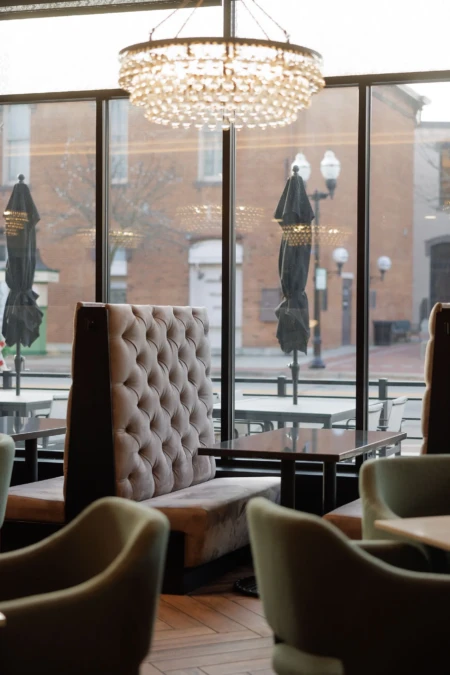 Luxurious restaurant interior with elegant chandelier, tufted seating, and large windows offering a street view. Outdoor umbrellas and lamps visible outside.