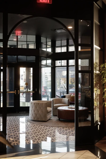 A modern lobby with comfortable seating, geometric flooring, and large glass doors. Illuminated "Exit" sign overhead, natural light streaming through windows.