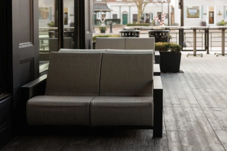 Outdoor seating area with cushioned benches on a wooden floor, overlooking a quaint town street with white buildings and decorative poles.