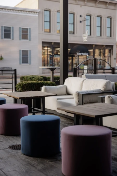 Modern outdoor seating area with cushioned benches and colorful stools. Background shows a storefront in a historic-style building, no recognizable landmarks.