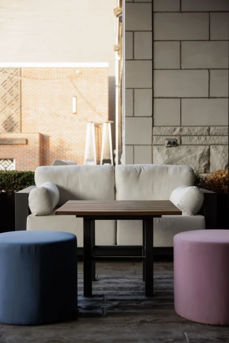 Cozy outdoor patio with a white sofa, wooden table, and colorful ottomans. Brick wall and geometric design in the background.