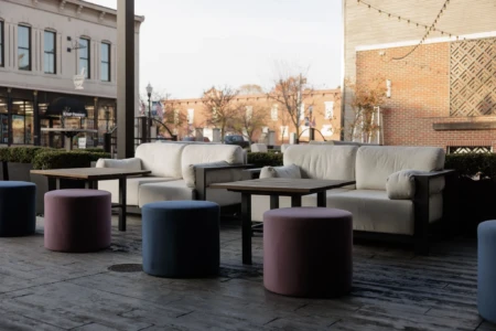 Outdoor patio with white sofas and colorful stools, overlooking a small-town street with storefronts, trees, and decorative lights. Empty seating area.