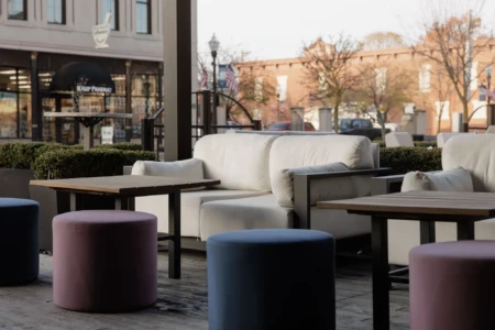 Outdoor seating area with cushioned chairs and tables, surrounded by colorful stools. Nearby, a pharmacy storefront and vintage-style buildings are visible.