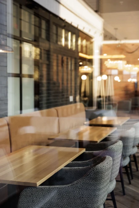 Empty modern café interior with wooden tables, cushioned chairs, and warm lighting, reflecting cozy ambiance through large glass windows.