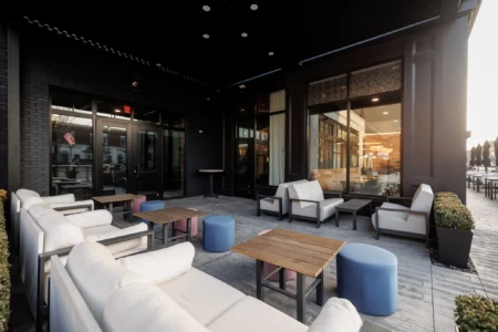 Outdoor patio with white sofas, wooden tables, and blue stools. Large glass windows reflect a building. No people are present. Cozy ambiance.