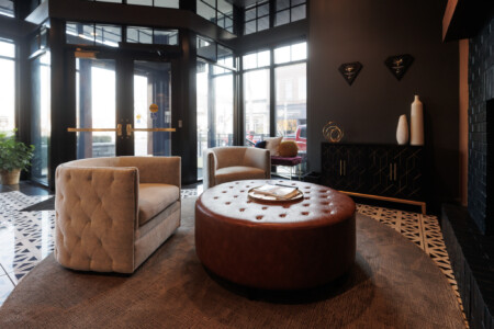Modern hotel lobby featuring stylish tufted armchairs, a round leather ottoman, and large windows allowing natural light. The decor includes geometric patterns on the floor and contemporary wall art, creating a welcoming atmosphere.