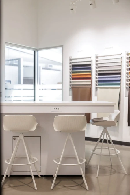 Modern design studio interior featuring a sleek white countertop and stylish bar stools, with a color wall displaying various fabric swatches for interior design. Natural light enters through large windows, enhancing the contemporary atmosphere.
