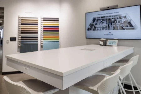 Modern design studio featuring a sleek white table with electrical outlets, surrounded by stylish white chairs. In the background, a wall displays a variety of fabric swatches in multiple colors, alongside a large screen showcasing a layout of the studio space.