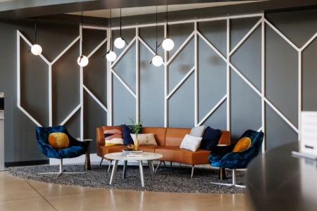 Modern lounge area featuring a stylish brown leather sofa and blue accent chairs, complemented by decorative pillows and a round coffee table, set against a geometric patterned wall and ambient pendant lighting.