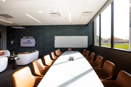 Modern conference room featuring a long white table surrounded by brown chairs, a large screen displaying the Midmark logo, and a whiteboard on a textured blue wall. Sunlight streams through large windows, illuminating the space and offering a view of the outdoor greenery.