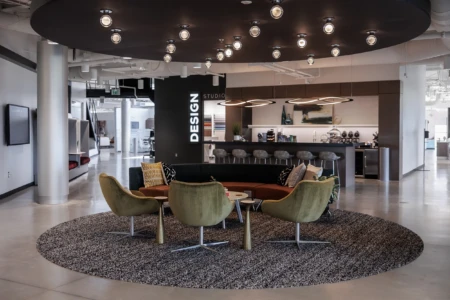 Modern design studio interior featuring a circular seating arrangement with green and gray chairs, a round coffee table, and a textured rug. The space includes stylish lighting, a dark ceiling, and a kitchenette area with bar seating in the background, emphasizing a contemporary aesthetic.