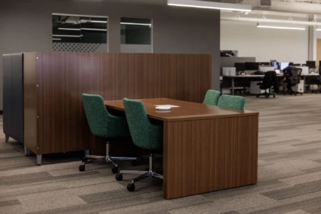 Modern office workspace featuring a wooden table and green upholstered chairs, separated by a wooden partition. The background shows additional workstations with computers, emphasizing a collaborative environment.